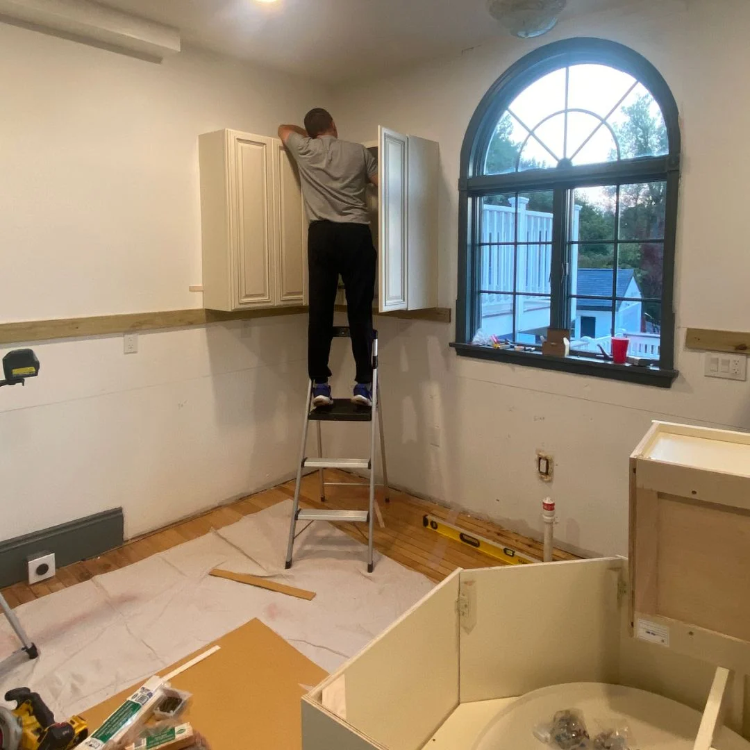 Person standing on a ladder installing kitchen cabinets in a home renovation setting, with tools and materials visible in the foreground, emphasizing quality craftsmanship in residential painting and remodeling services.